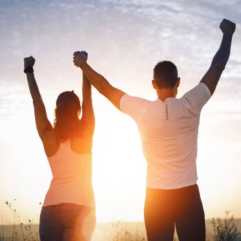 A man and woman holding hands above their head with the sunset in front of them.