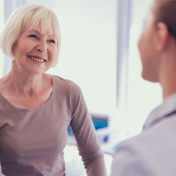 An older woman in a tan long sleeved shirt smiling while speaking to a provider with a plain gray green background.