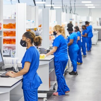 A row of technicians in blue scrubs working at individual work stations.