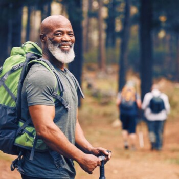 An African American man with a gray hair wearing a green backpack and gray shirt hiking in the woods on a trail.
