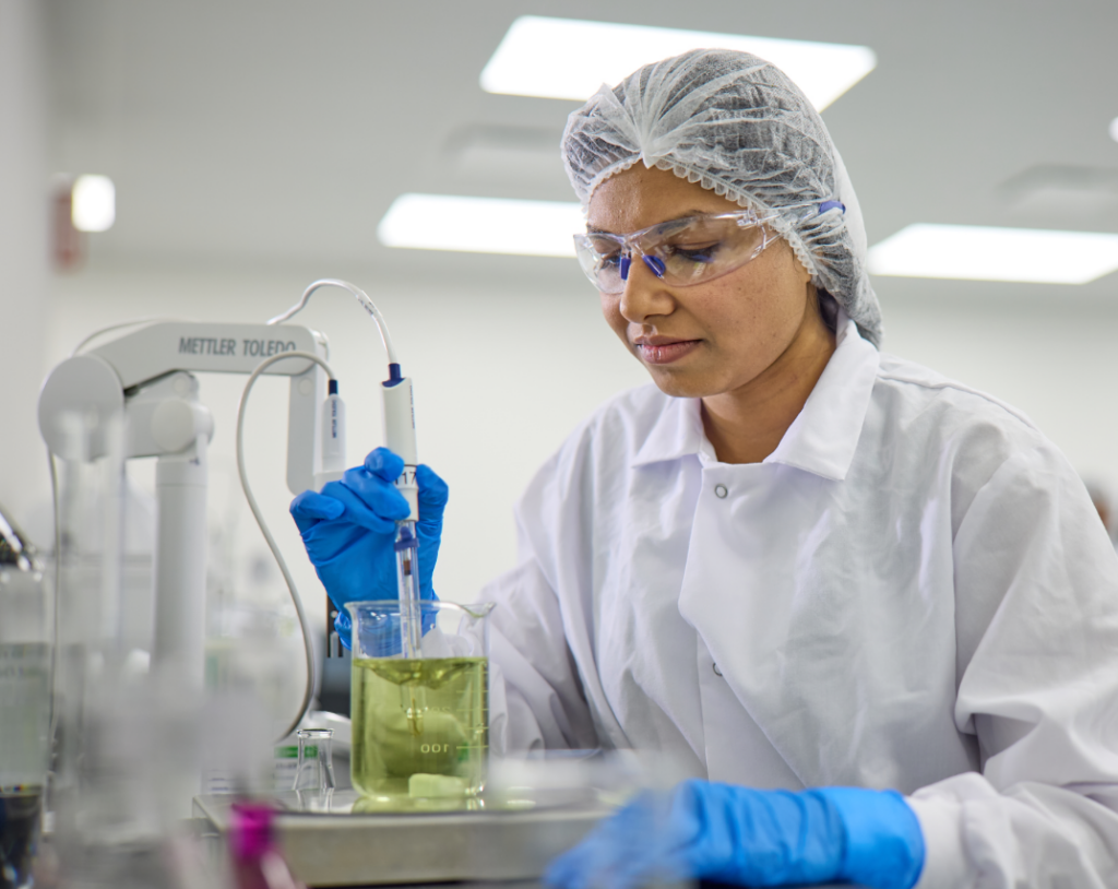 A woman in a white clean suit using a machine in a beaker of colored liquid.