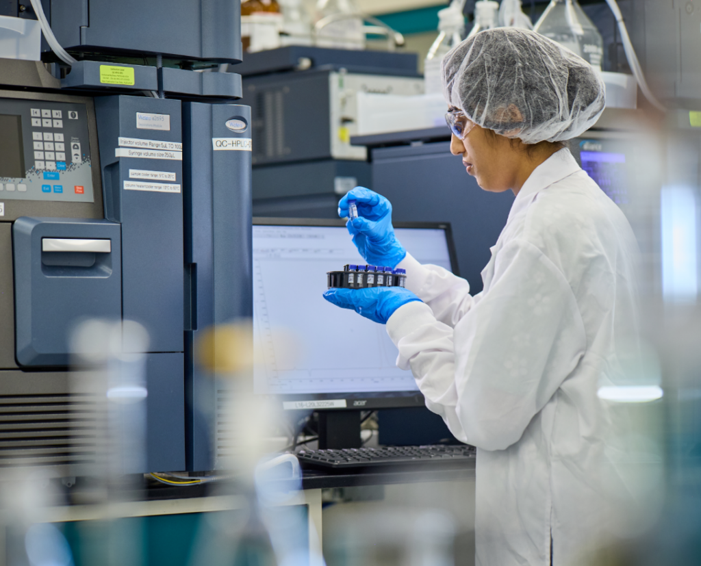 A woman in a white clean suit and a hairnet holding a vial in a gloved hand.