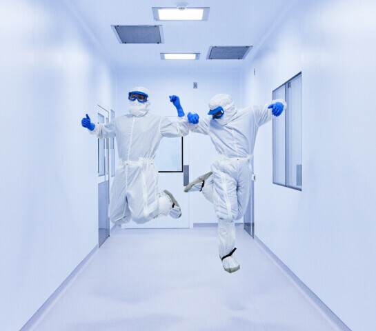 Two workers in white clean suits, goggles, and blue gloves jumping in the air.