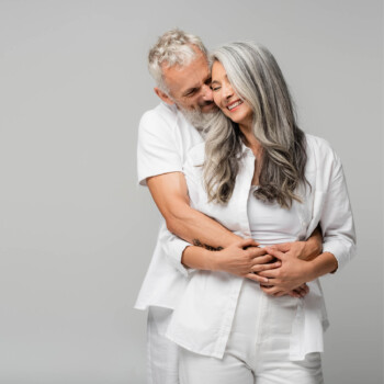 A older man hugging a smiling older woman from behind both wearing white